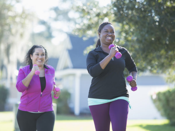 两个多民族妇女在户外锻炼，在一个阳光明媚的日子里，在一个居民区慢跑，手里拿着哑铃。影片的主要焦点是那位非裔美国妇女，当她的西班牙朋友试图赶上她时，她笑了。