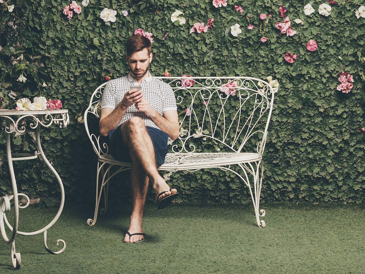 英俊的男子坐在老式的长凳上打字短信，绿色的墙壁上的树叶和花朵的背景。