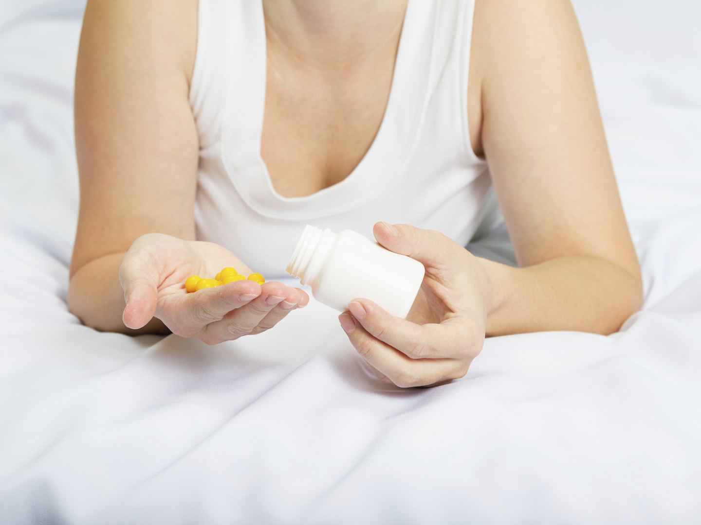 年轻的白人女人穿着睡衣躺在床上，手里拿着白色瓶子里的黄色药片。