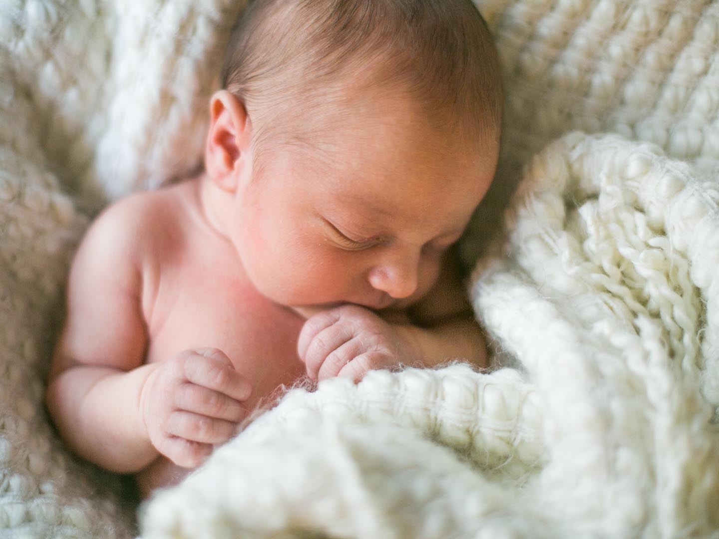 一个刚出生一周的男婴裹着白色毛茸茸的毯子睡觉。他很平静，他的手放在嘴边。