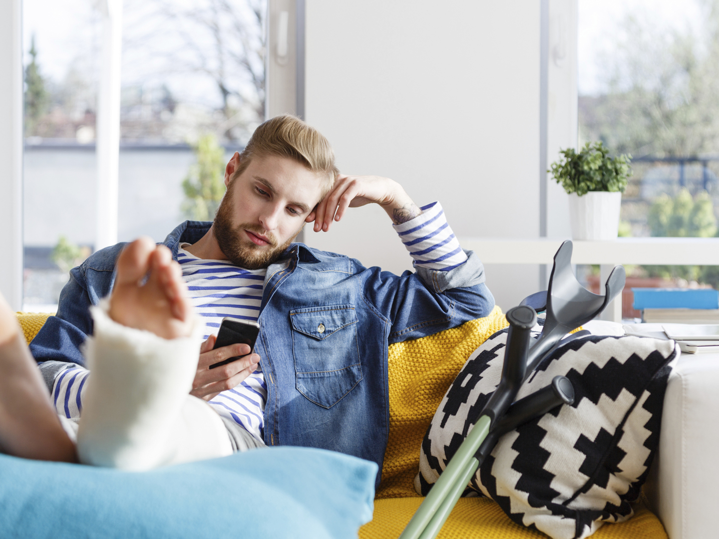年轻男子断腿石膏石膏躺在沙发上，在家里使用智能手机。