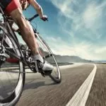 一个运动员在路上骑自行车。该男子身穿黑色自行车短裤和护腿，红色无袖上衣，戴着红白相间的头盔和太阳镜。图像在运动中变得模糊。
