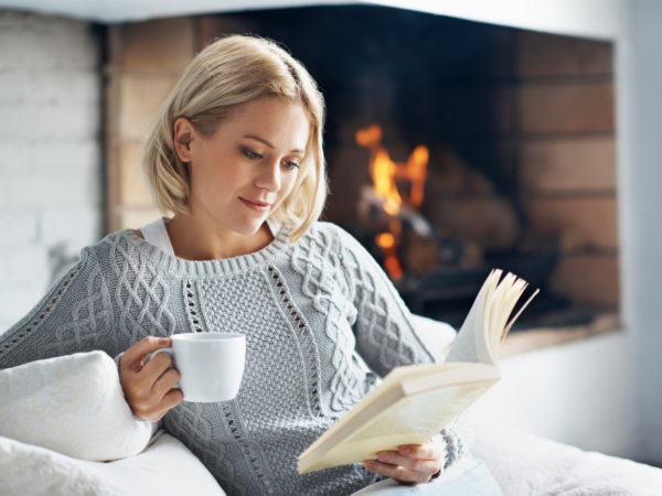 一个美丽的年轻女子在壁炉边看书，喝着热饮