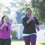 两个多民族妇女在户外锻炼，在一个阳光明媚的日子里，在一个居民区慢跑，手里拿着哑铃。影片的主要焦点是那位非裔美国妇女，当她的西班牙朋友试图赶上她时，她笑了。